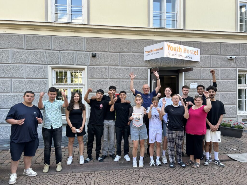 Eine Gruppe von Jugendlichen stehen mit ihrem Betreuer vor dem "Youth Hostel" in Brixen. Sie lächeln und jubeln. Eine Jugendliche hält das BSJ Logo auf einem Plastik-Wappen in der Hand.