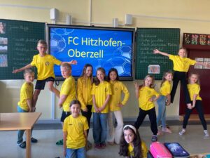 Kinder in gelben Trikots stehen im Klassenzimmer vor ihrer Tafel, auf der der Vereinsname des FC Hitzhofen Oberzell eingeblendet ist.