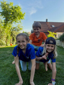 Drei strahlende Kinder des TSV Mainburg in Vereinsshirts knieen im Garten.