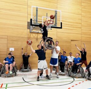 Die Trainer des USC München heben einen Spieler mit seinem Rollstuhl für einen Spaß-Dunking in die Höhe zum Korb.