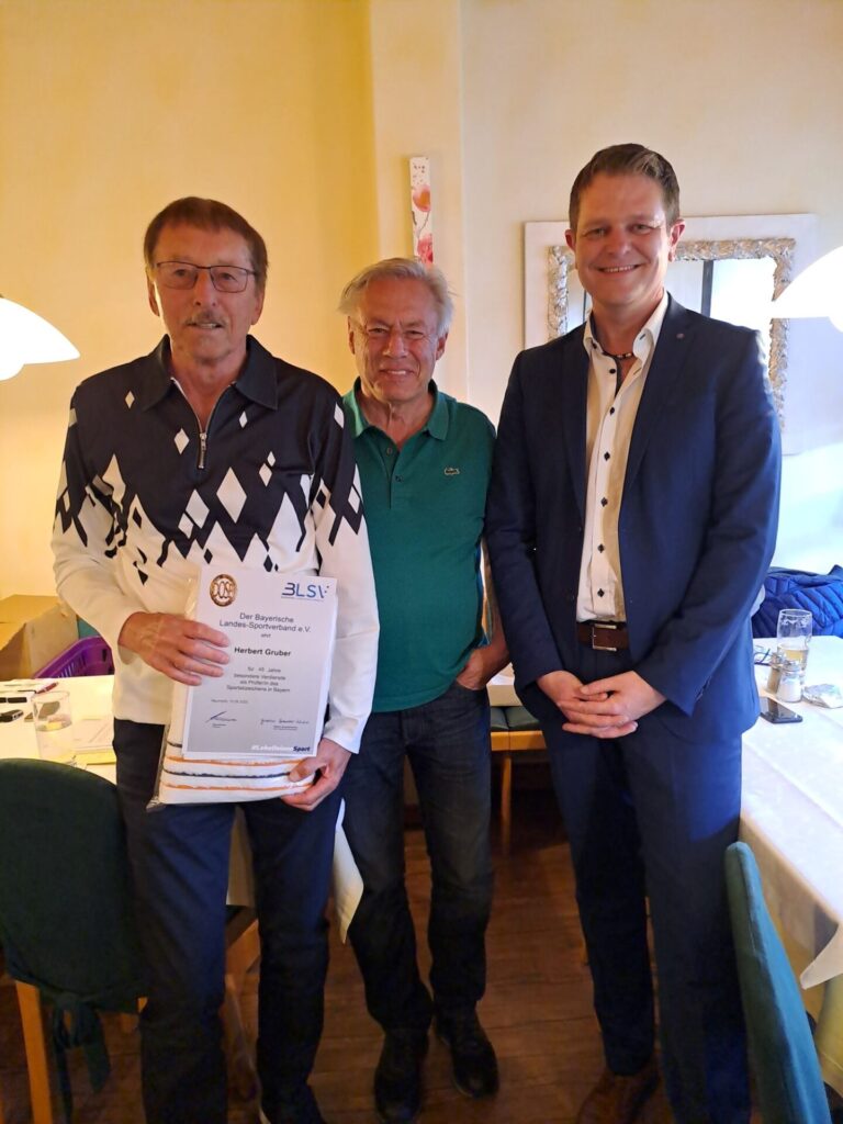 Drei Personen lächeln für ein Foto in die Kamera. Links befindet sich Herbert Gruber, der eine Urkunde in der Hand hält.