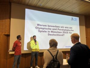 Zwei Männer sprechen vor einer Leinwand mit der Aufschrift "Warum bewerben wir uns um Olympiasche und Paralympische Spiele in München 20XX für Deutschland?"