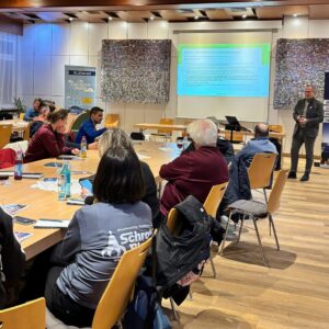 Mehrere Menschen sitzen in einem Raum und schenken der vortragenden Person ihre Aufmerksamkeit