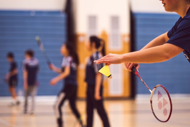 Mehrere Personen stehen in einer Sporthalle. Von der vordersten Person ist nur der Arm und der Badmintonschläger sowie -ball zu sehen. Die anderen Personen im Hintergrund sind nur verschwommen wahrzunehmen.