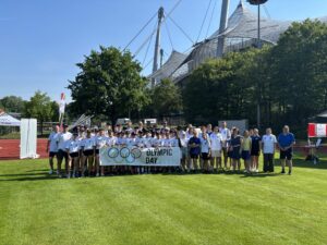 Ein Gruppe von Menschen steht auf einer Wiese vor dem Olympiastadion in München und hält ein Schild mit der Aufschrift "Olympic Day"